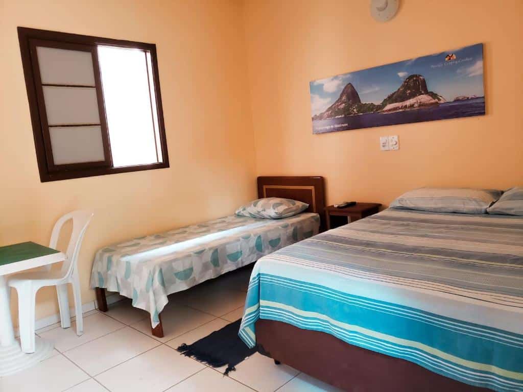 Quarto da Pousada Camping Camburi com uma cama da solteiro do lado esquerdo e uma cama de casal lado direito uma mesa com uma cadeira de plástico branca em frente a cama de solteiro.