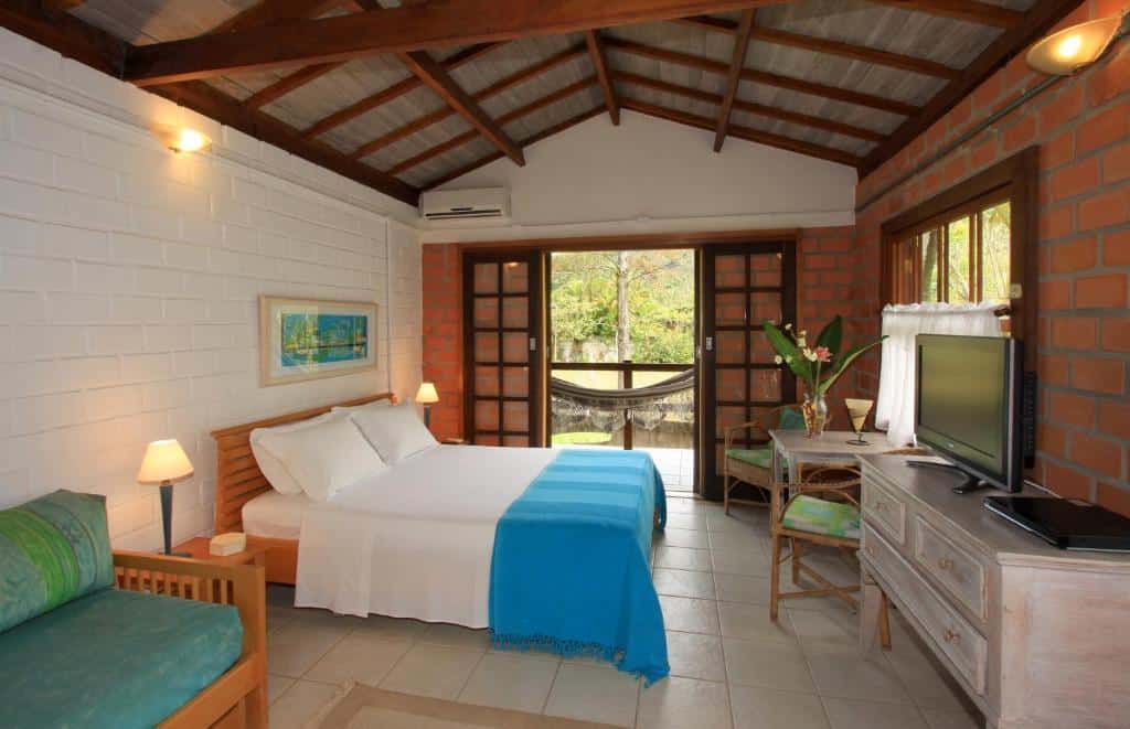 Quarto da Pousada do Rosa Camburi com cama no centro do quarto, sofá do lado direito, em frente a cama tem uma cômoda com TV e mesa com duas cadeiras. Do lado esquerdo, porta ampla de madeira com sacada com rede.
