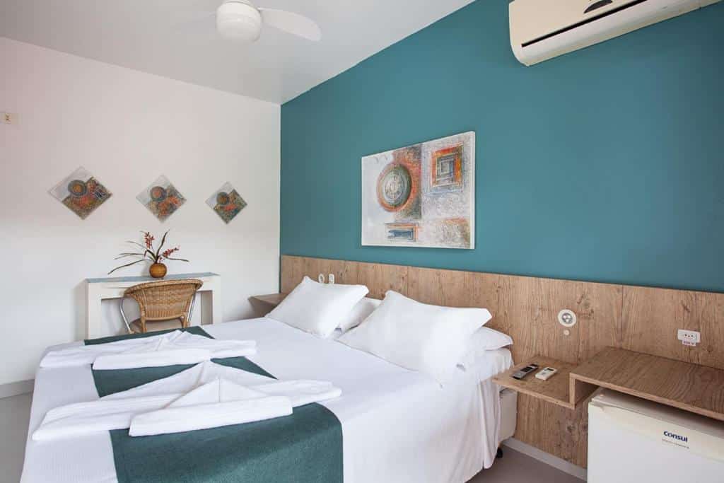 Quarto da Camburioca com cama de casal no centro do quarto, do lado direito da cama frigobar e do lado esquerdo mesa de trabalho branca com cadeira de palha e um vaso de planta em cima da mesa.