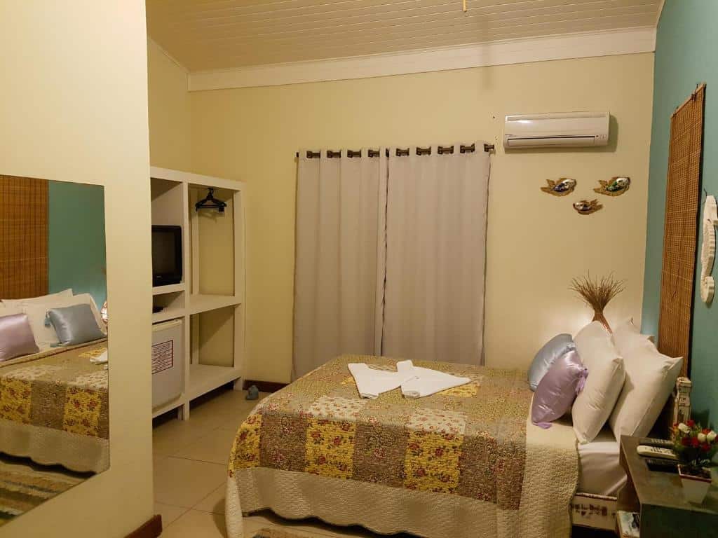 Quarto da Coco Bambu Cambury com espelho no lado esquerdo do quarto, cama de casal do lado do direito com duas cômodas ao lado da cama, ao fundo, janela com cortinas.