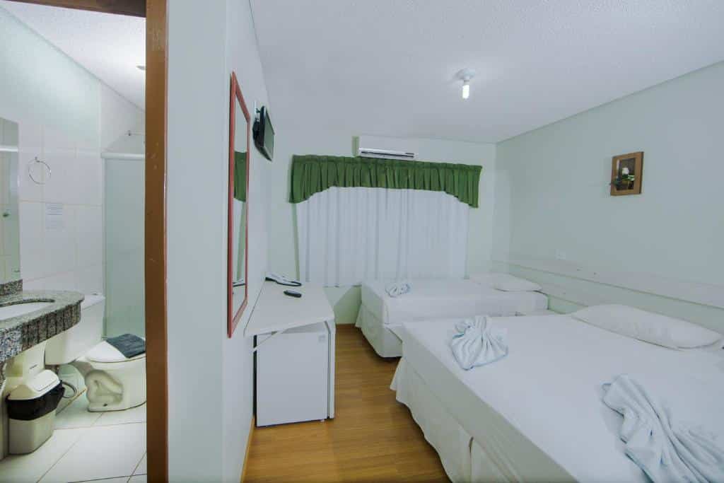 Quarto da Pousada Caroline que mostra do lado direito o quarto com uma cama de casal, uma cama de solteiro, frigobar, tv. Já do lado esquerdo separado por uma parede tem o banheiro.