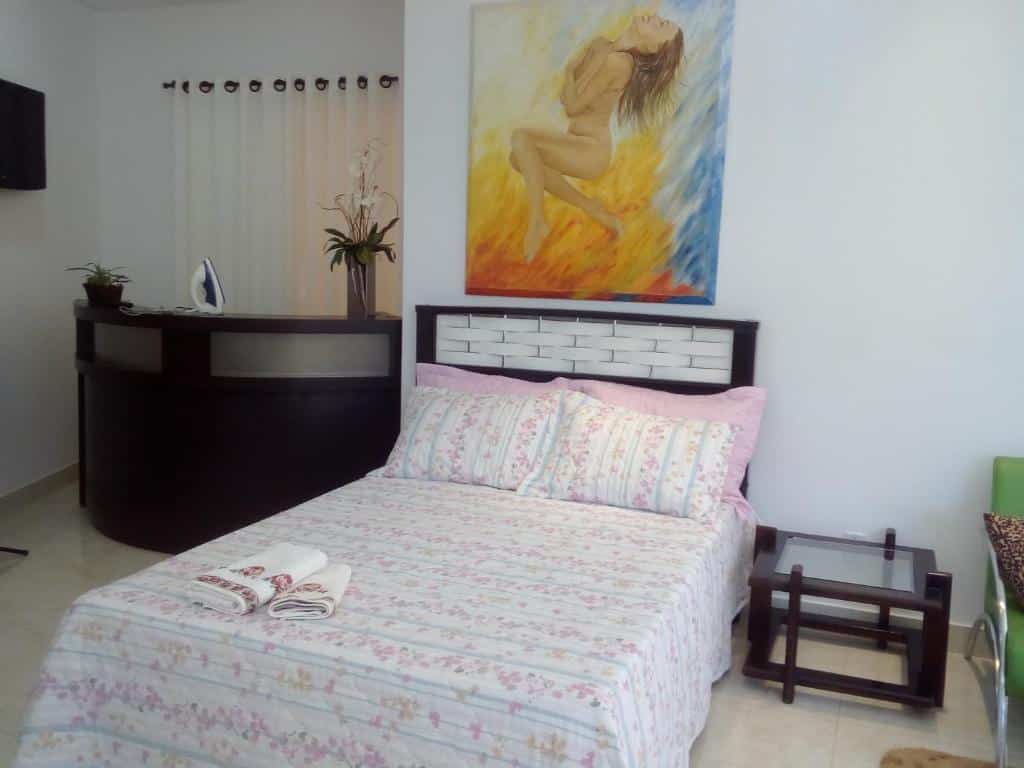 Quarto da Pousada Bom Sono com uma cama de casal e quadro enorme na parede. Do lado direito da cama tem uma mesinha de vidro e uma cadeira, já do lado esquerdo da cama tem uma bancada com alguns objetos, uma cortina e uma tv na parede.
