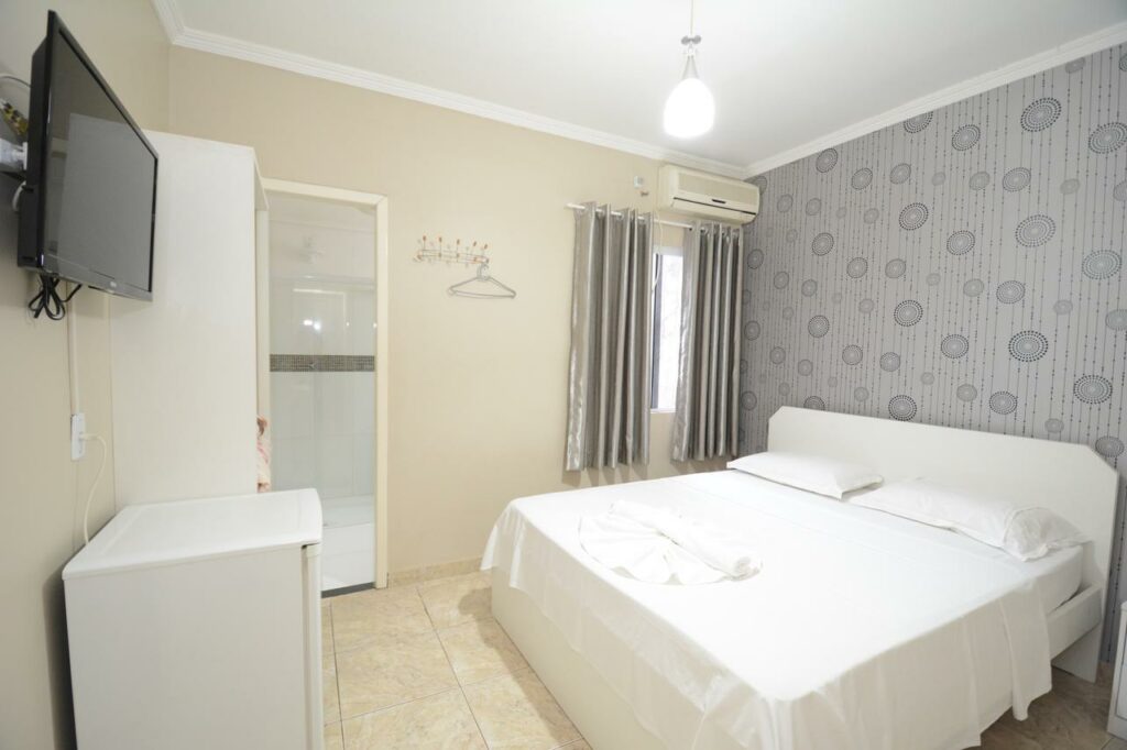 Quarto da Pousada Quedas D'água com uma cama de casal no centro do quarto e do lado esquerdo tem uma parede com uma janela, um ar-condicionado e do lado uma porta. Em frente a cama tem um guarda-roupa, um frigobar e uma tv na parede.