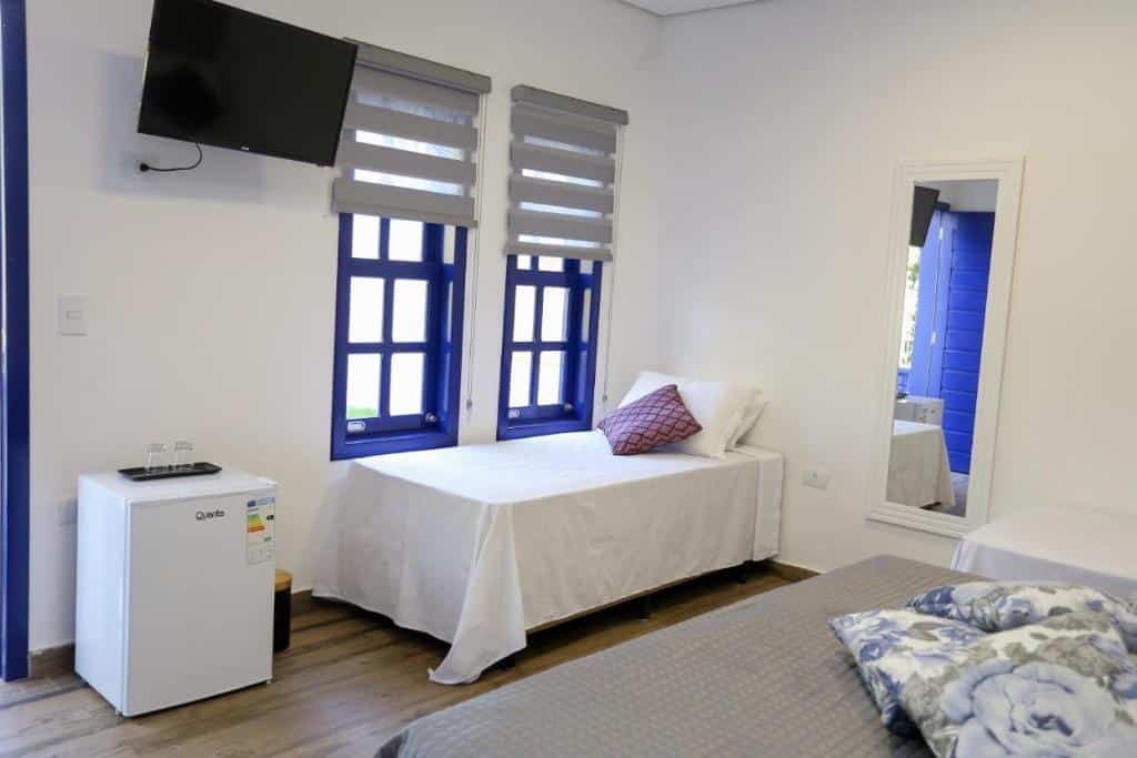 Quarto do Pequeno Paraíso com uma cama de casal e do lado uma de solteiro. Na frente das duas camas tem uma cama de solteiro do lado de duas janelas na parede e na frente um frigobar e uma tv na parede.