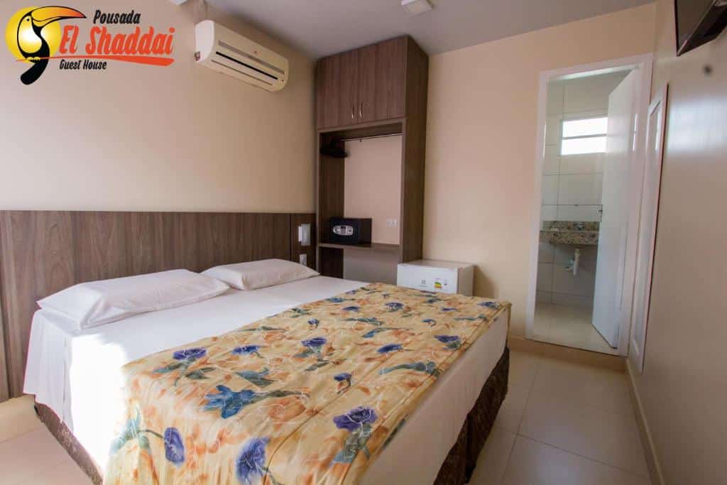 Quarto da Pousada El Shaddai que mostra uma cama de casal e do lado esquerdo da cama um guarda-roupa, do lado um frigobar e uma porta que dá acesso ao banheiro. No quarto há também um ar-condicionado e uma tv na parede de frente pra cama.