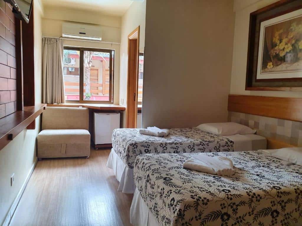 Quarto da Pousada Sonho Meu Foz com duas camas de solteiro do lado direito e um quadro na parede. Já do lado esquerdo tem uma tv na parede ao fundo uma poltrona, um frigobar, um ar-condicionado e uma janela de vidro.