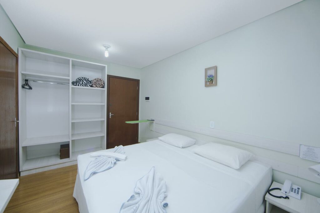 Quarto do Pousada Iguassu Charm Suites que mostra uma cama de casal com uma mesinha do lado da cama com um telefone. Já do lado esquerdo da imagem tem um guarda-roupa e de cada lado dele tem uma porta de madeira.