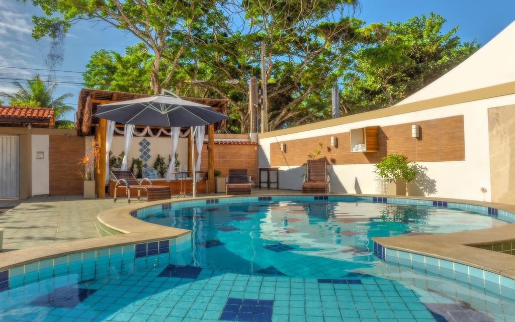 Piscina da pousada Lua e Mar em Ilhéus, com espreguiçadeiras, um pergolado e um portão e árvores ao fundo