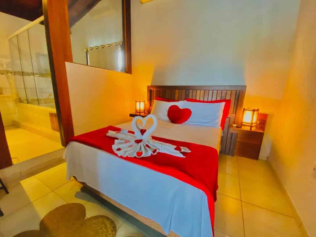 Uma cama na Pousada Gauleses com roupa de cama vermelha, um travesseiro de coração e cisnes de toalha está em um quarto bem iluminado ao lado de um banheiro.