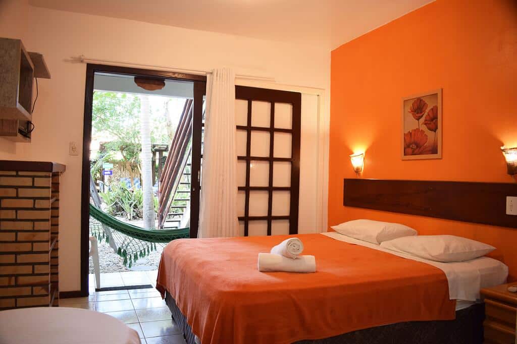 Um quarto aconchegante da Vila dos Coqueiros com uma parede laranja de destaque, cama de casal, toalhas enroladas e uma rede do lado de fora da porta de vidro deslizante.