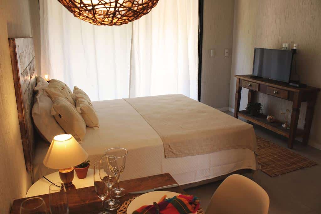 Um quarto da Pousada Vivenda do Rosa com uma cama de casal, abajur de mesa de cabeceira, TV em um suporte de madeira e uma pequena mesa de jantar em primeiro plano.