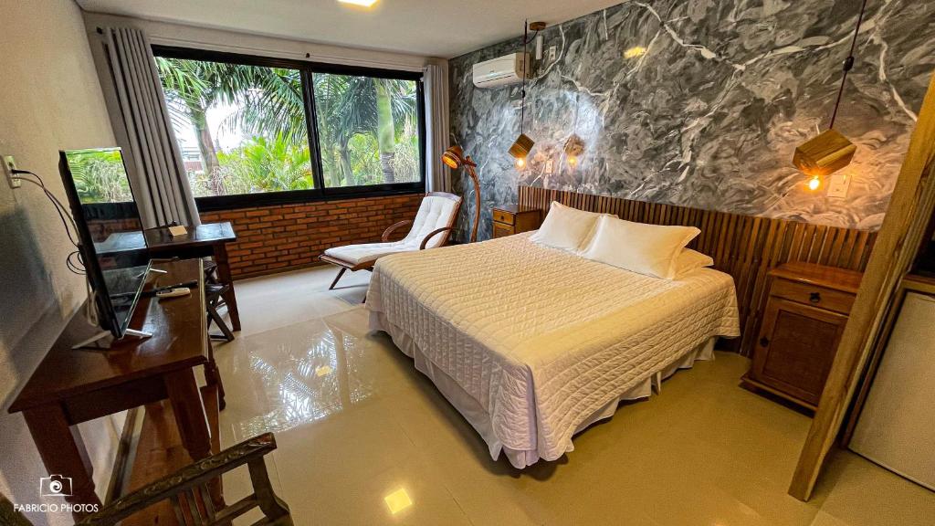 Uma cama bem arrumada em um quarto do Pousada Descanso do Rei com uma parede de mármore, escrivaninha, poltrona e uma grande janela com vista.