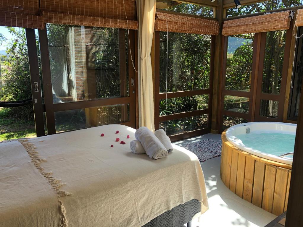 Uma cama da Pousada Watu Kerere com toalhas e pétalas de rosas ao lado de uma banheira de hidromassagem redonda em uma sala iluminada pelo sol com grandes janelas e vista para o jardim.