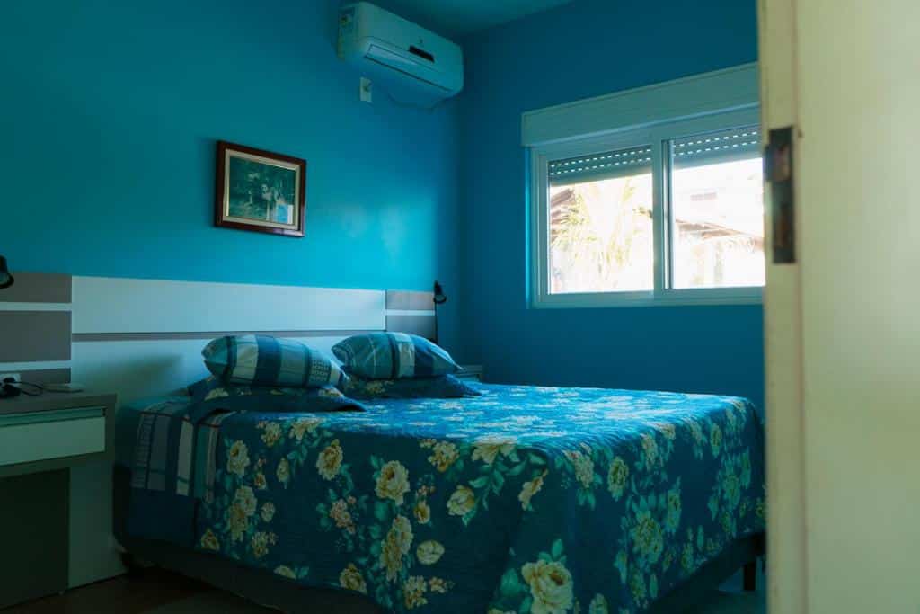 Um quarto da Pousada Praia da Villa com paredes azuis, uma colcha floral, dois travesseiros, uma janela e um ar-condicionado na parede.