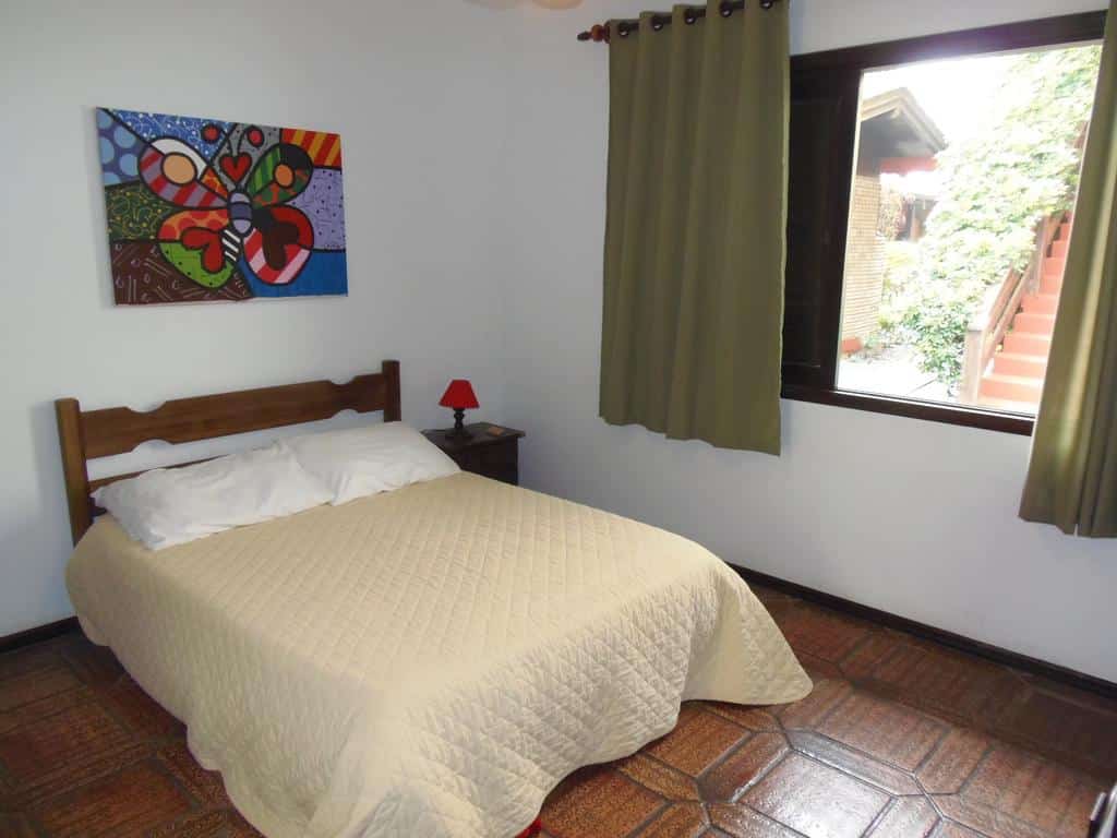 Uma cama bem arrumada na Pousada da Villa com uma colcha bege, uma mesinha de cabeceira, um abajur vermelho, uma janela com cortinas verdes e arte colorida na parede.