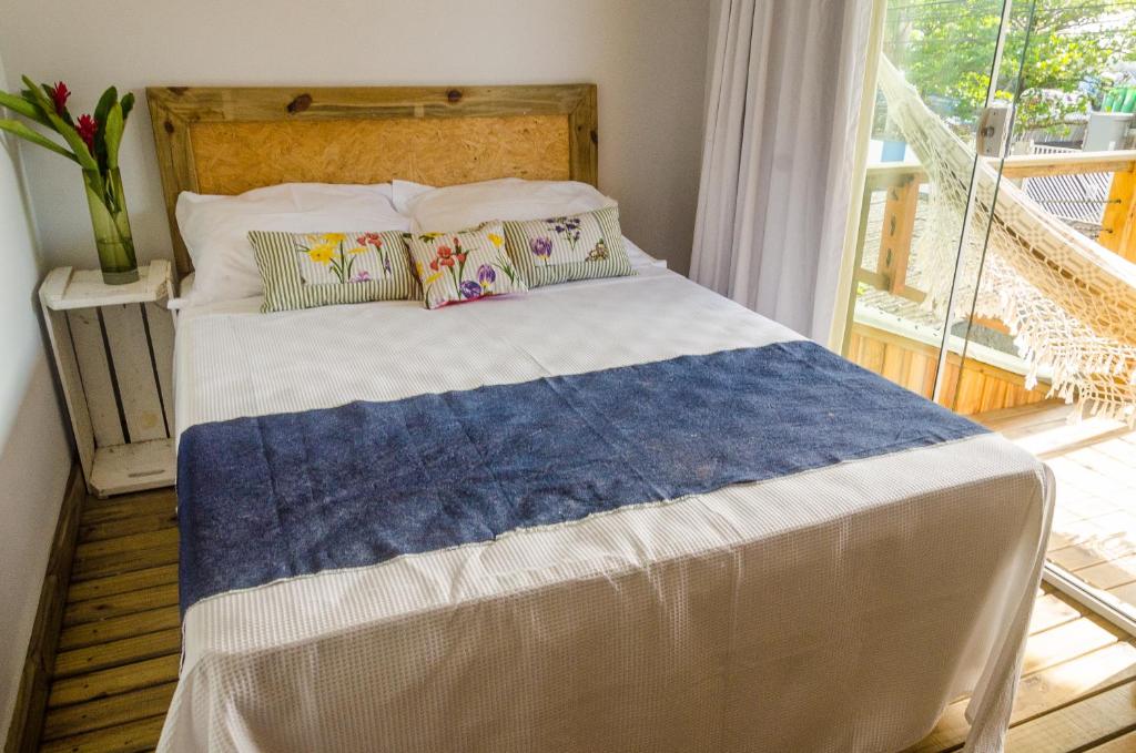 Uma cama bem arrumada com travesseiros florais e um tapete azul fica ao lado de uma janela com uma rede e vegetação do lado de fora, no Alma Brava, para representar pousadas em Itajaí.