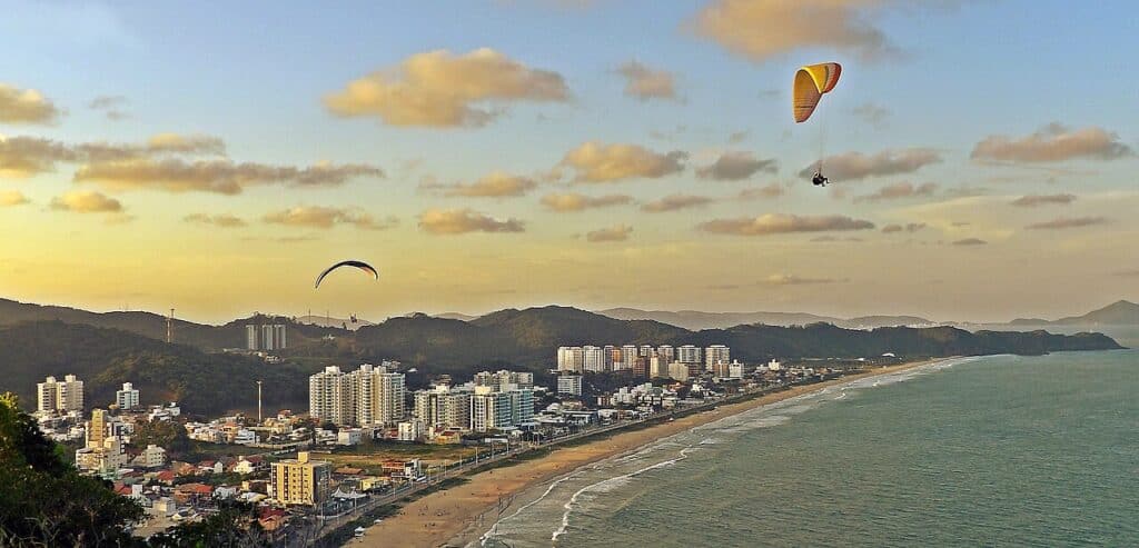Cidade litorânea de Itajaí com prédios altos, praia de areia, oceano, colinas distantes e duas pessoas voando de parapente ao pôr do sol.