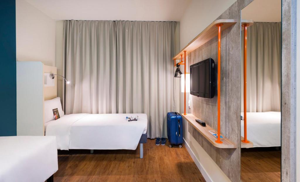 Um quarto no ibis Budget Navagantes Itajaí com uma cama de solteiro, uma TV na parede, uma mala azul e cortinas cinza cobrindo a janela, para representar pousadas e hotéis em Itajaí.