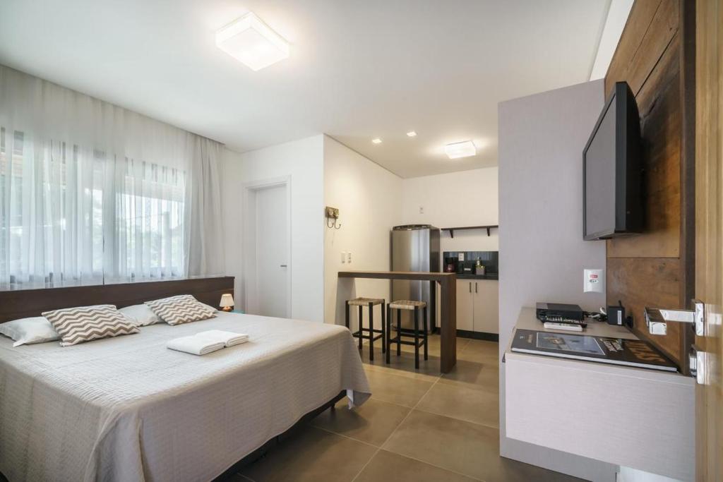 Estúdio moderno com uma cama, pequena área de cozinha, balcão com duas banquetas, TV e janela grande com cortinas transparentes, no Banzai Brava Suítes, para representar pousadas em Itajaí.