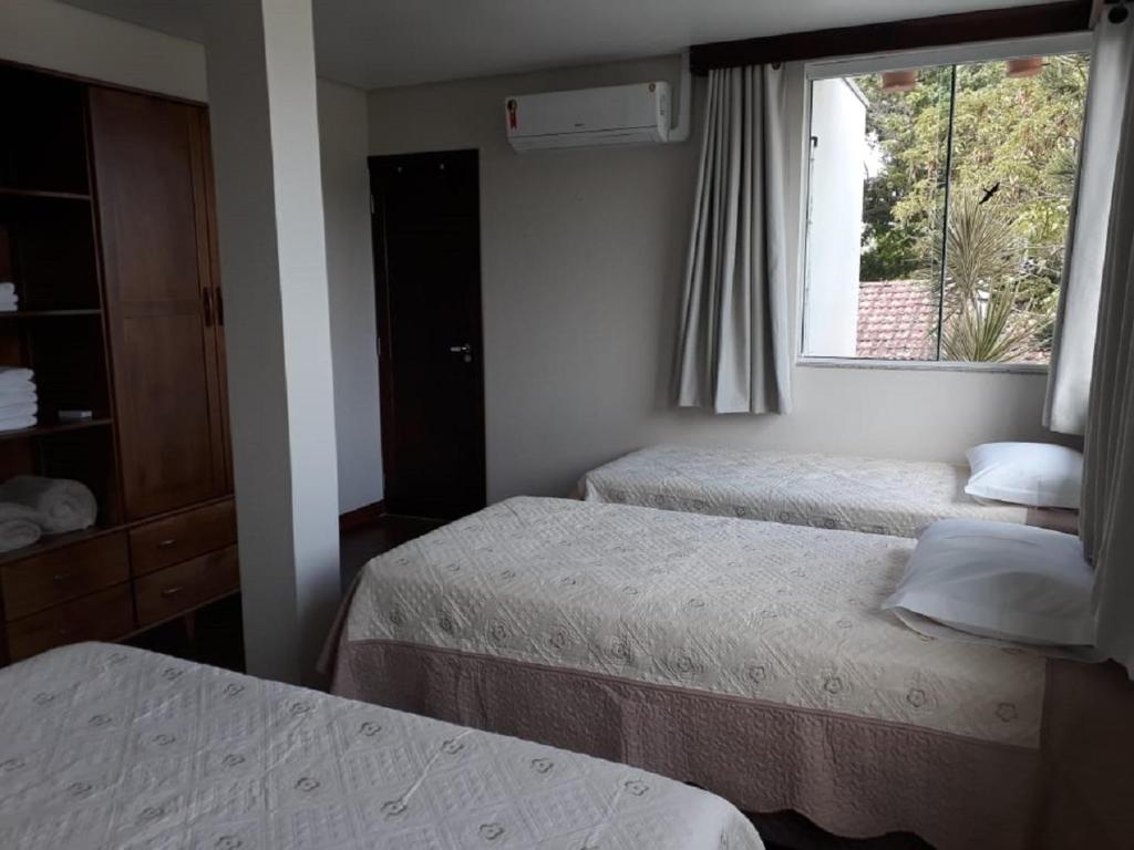 Três camas de solteiro com edredons brancos em um quarto com janela, ar-condicionado e guarda-roupa de madeira, na Casa da Praia, para representar pousadas em Itajaí.