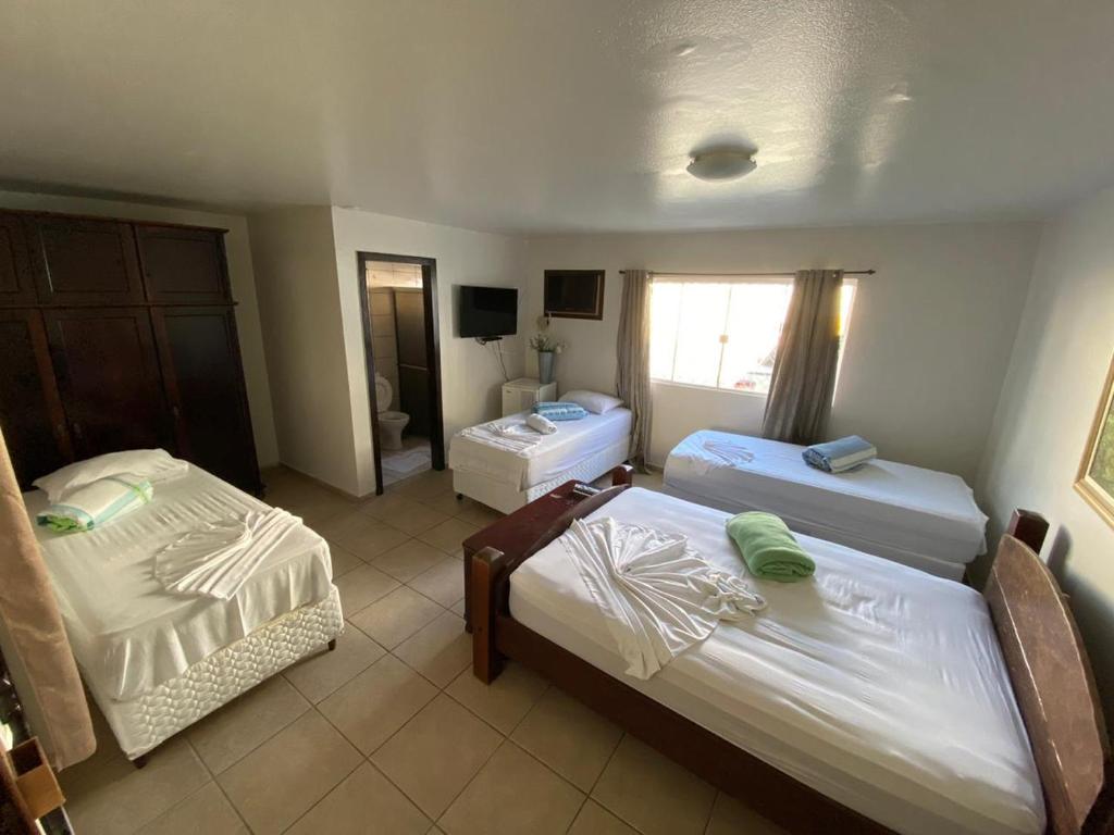 Quarto no Hotel Roda do Mar Inn Itajaí Navegantes com três camas de solteiro, toalhas dobradas, um guarda-roupa, piso de azulejo, janela com cortinas e uma TV na parede, para representar pousadas em Itajaí.