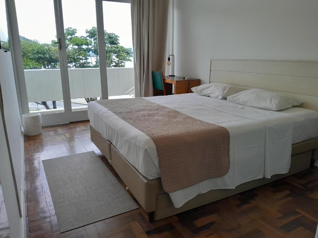 Uma cama bem arrumada em um quarto bem iluminado com piso de madeira, uma pequena escrivaninha e portas de vidro que dão para uma varanda com árvores do lado de fora, no Hotel Marambaia Cabeçudas - frente mar, para representar pousadas em Itajaí.