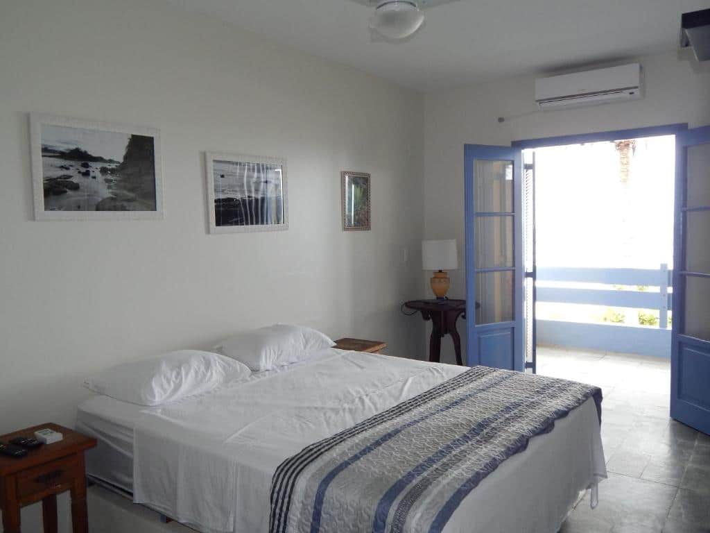 Um quarto simples da Pousada Sobre as Pedras com uma cama de casal, duas mesinhas de cabeceira, fotos emolduradas na parede e uma porta aberta para uma varanda.