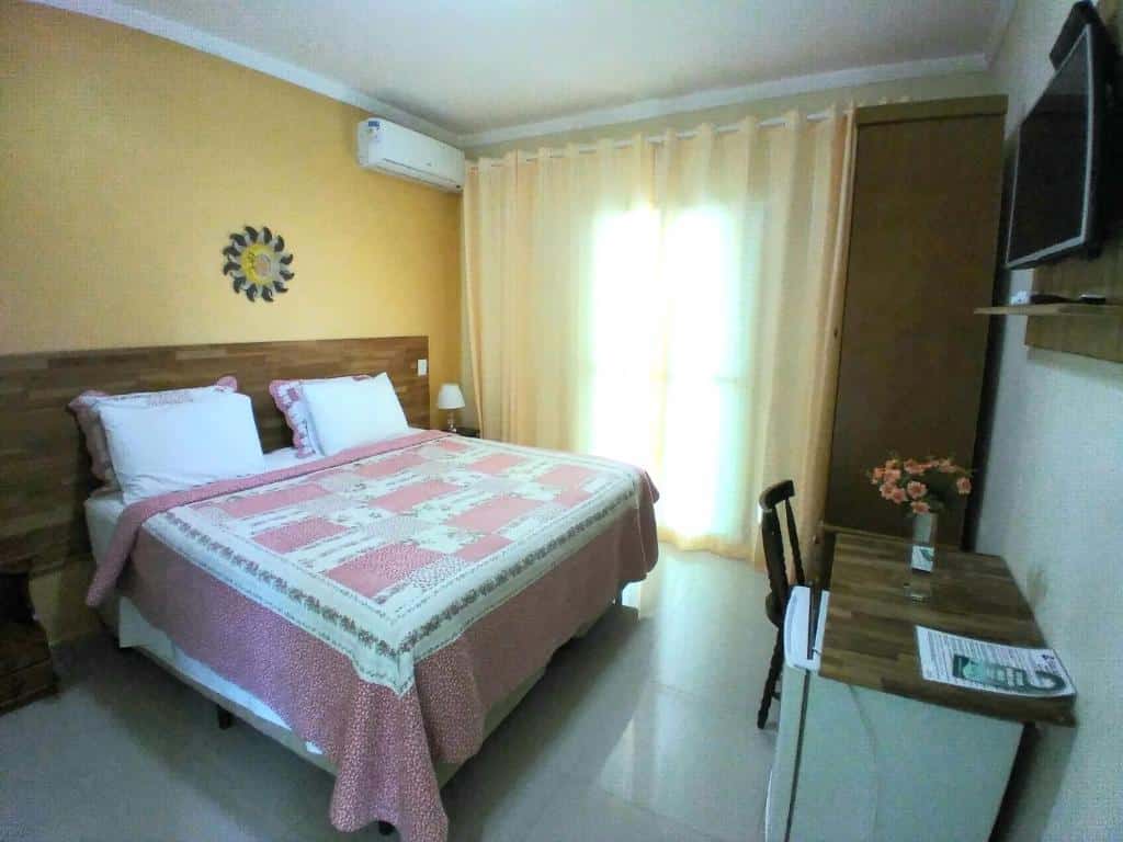 Um pequeno quarto da Visual Mar Pousada com uma cama de casal, uma TV na parede, uma escrivaninha, uma cadeira e uma janela com cortinas transparentes.