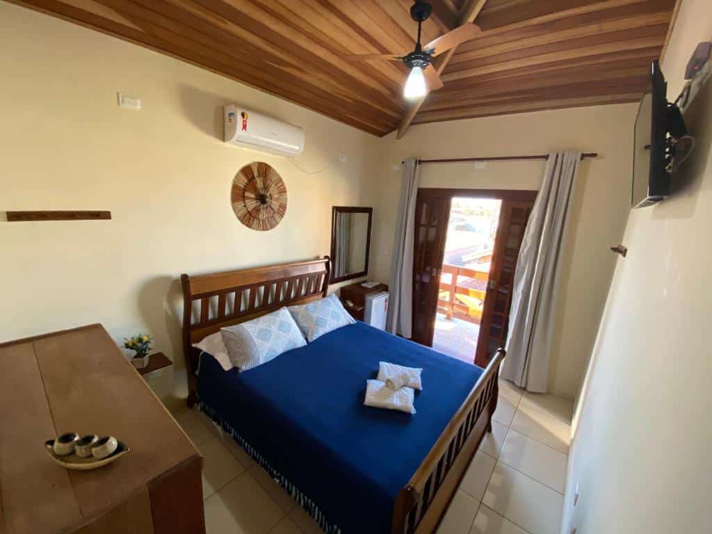 Uma cama bem arrumada com roupa de cama azul em um pequeno quarto da Pousada da Brisa com teto de madeira, cômoda, TV e acesso à varanda.