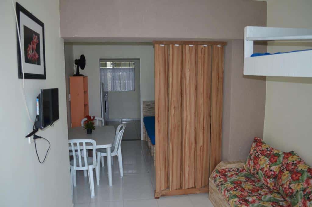 Pequena sala de estar da Pousada/Flat Praia Do Sonho com um sofá floral, mesa de jantar, TV na parede, beliche e divisória de madeira separando as áreas.