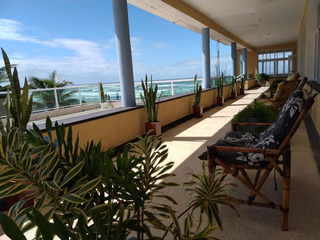 Uma varanda longa e coberta do Hotel Pousada Miami com vasos de plantas, cadeiras almofadadas e vista para o oceano sob um céu parcialmente nublado.