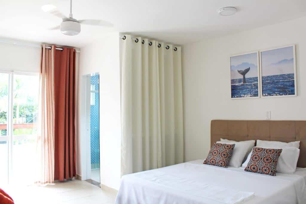Um quarto bem iluminado da Murakami Pousada com uma cama de casal, travesseiros, arte na parede, ventilador de teto, cortinas e portas de vidro deslizantes.