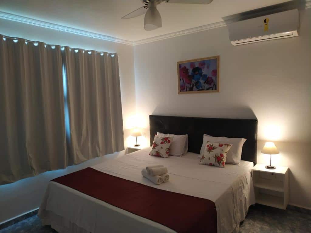 Uma cama bem arrumada na Pousada Elegance Beira Mar com roupas de cama brancas e detalhes em vermelho, ladeada por duas mesinhas de cabeceira com abajures, sob um ar-condicionado montado.