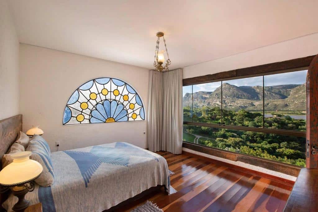 quarto da pousada Sítio Jatoba na Lapinha da Serra com uma cama de casal à esquerda, um vitral de flores azul, branca e amarela na parede ao fundo e uma ampla janela de vidro com vista para a Serra do Cipó mostrando várias árvores, um lago e lindas montanhas.