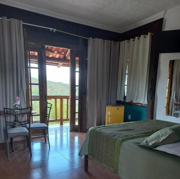 interior do quarto na Pousada Cachoeiras do Cipó na Lapinha da Serra com uma cama de casal à direita, uma mesa de vidro com duas cadeiras à esquerda e uma varanda com vista para a serra ao fundo.