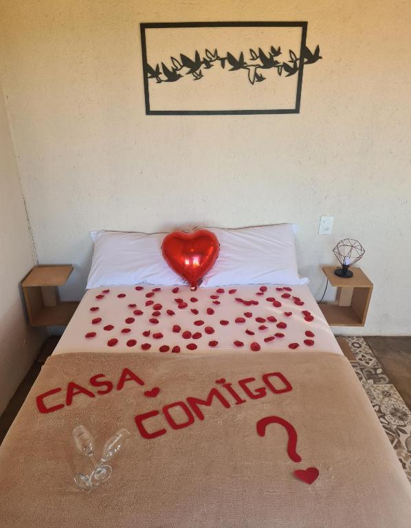 cama de casal enfeitada com pétalas de rosa vermelhas formando os dizeres "casa comigo?'. De cada lado da cama há uma pequena cômoda com um abajur e, acima da cama, há um quadro vazado de pássaros voando.