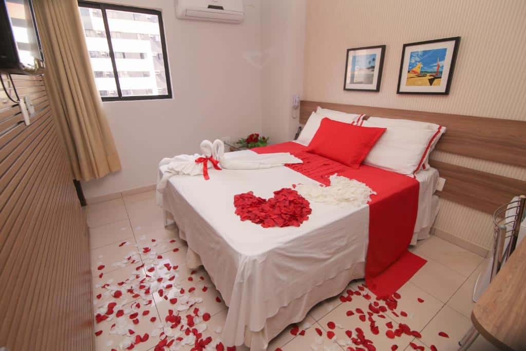 Quarto romântico da Pousada Capital das Águas com uma cama de casal com pétalas de rosas sob ela e o chão, além de uma televisão e um ar-condicionado, para representar pousadas em Maceió.