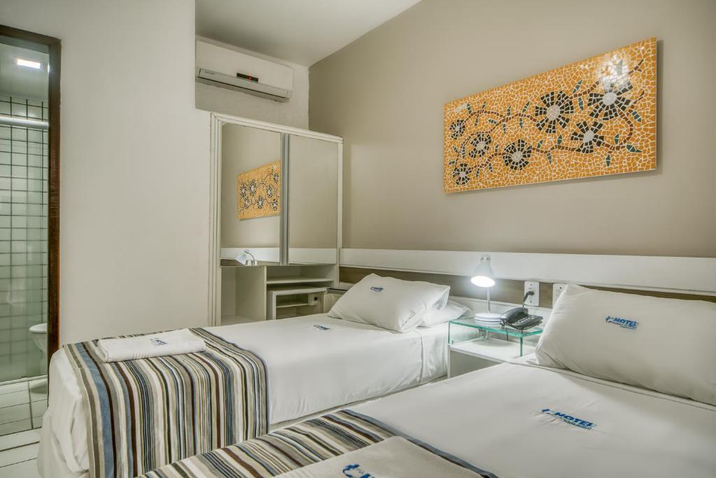 Duas camas de solteiro em um quarto na Pousada da Sereia na Praia de Pajuçara, com roupa de cama listrada, um espelho de parede, uma escrivaninha e obras de arte em mosaico acima das camas, para representar pousadas em Maceió.