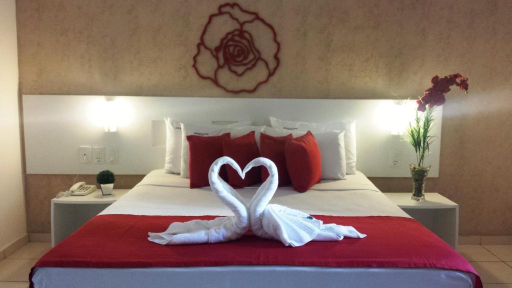 Quarto na Pousada Perola Dourada na Praia de Jatiúca, com uma cama, roupa de cama vermelha e branca, toalhas de cisne, travesseiros vermelhos, arte na parede, abajur de cabeceira e flores, para representar pousadas em Maceió.