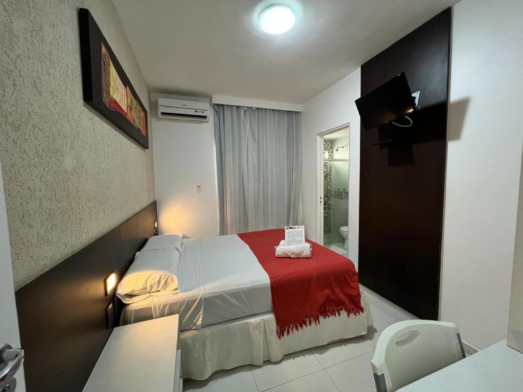 Um pequeno quarto na Pousada Aquarela do Brasil na Praia de Pajuçara, com uma cama de casal, cobertor vermelho, TV de parede, ar-condicionado e banheiro anexo, para representar pousadas em Maceió.