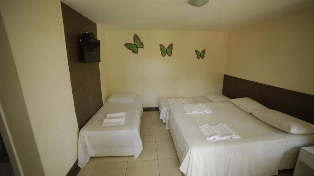 Um quarto na Pousada das Araras na Praia Cruz das Almas, com uma cama de solteiro e uma cama de casal, ambas com lençóis brancos; três decorações de borboletas na parede, para representar pousadas em Maceió.