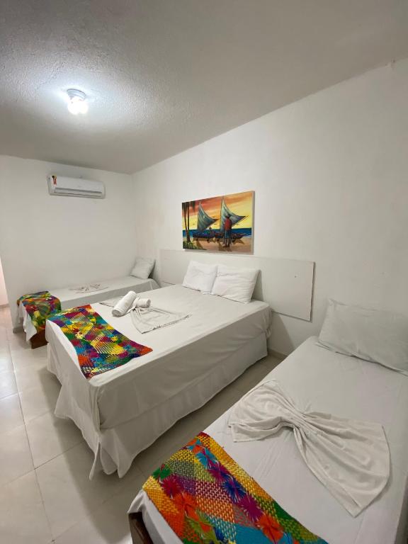 Um quarto na Pousada Nossa Casa na Praia de Jatiúca, com uma cama de casal e duas camas de solteiro, mantas coloridas, paredes brancas e um quadro acima da cabeceira da cama, para representar pousadas em Maceió.
