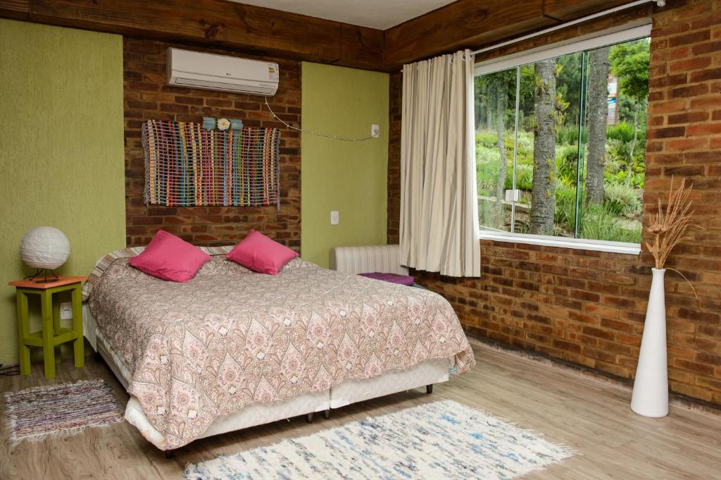 Quarto do Bina's Haus Pousada, Cafeteria e Atelier, uma das pousadas em Nova Petrópolis, com paredes de tijolos, uma cama de casal com roupa de cama estampada, dois travesseiros rosa e uma grande janela com vista para o jardim.