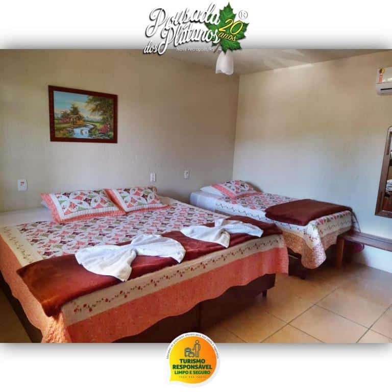 Um quarto da Pousada dos Plátanos com duas camas, um quadro na parede, toalhas nas camas e um ar-condicionado sobre um espelho.