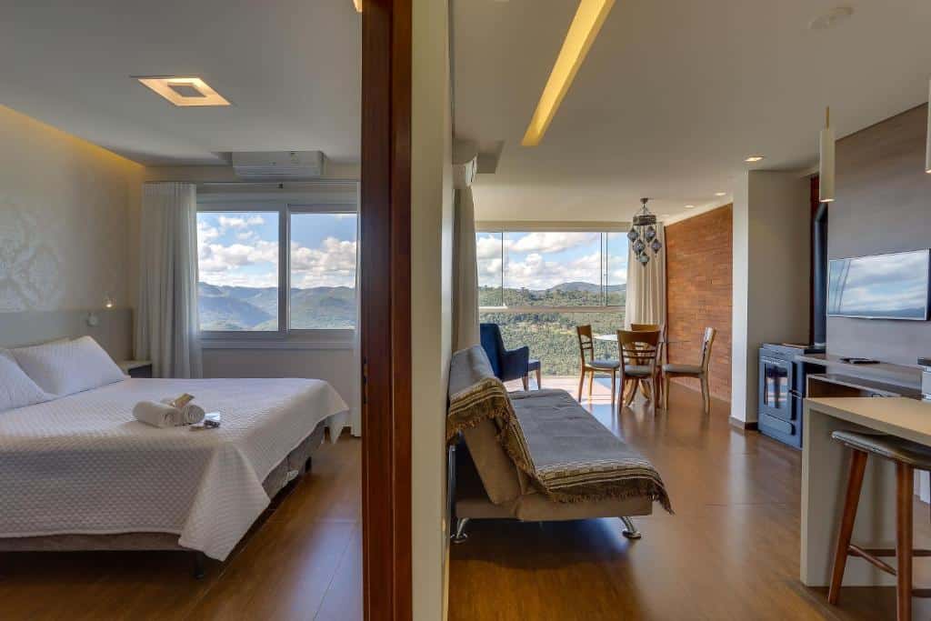 Uma moderna suíte da Pousada Janela do Vale, uma das pousadas em Nova Petrópolis, com cama, sofá, área de jantar, TV, janelas grandes e vista para a montanha sob um céu parcialmente nublado.