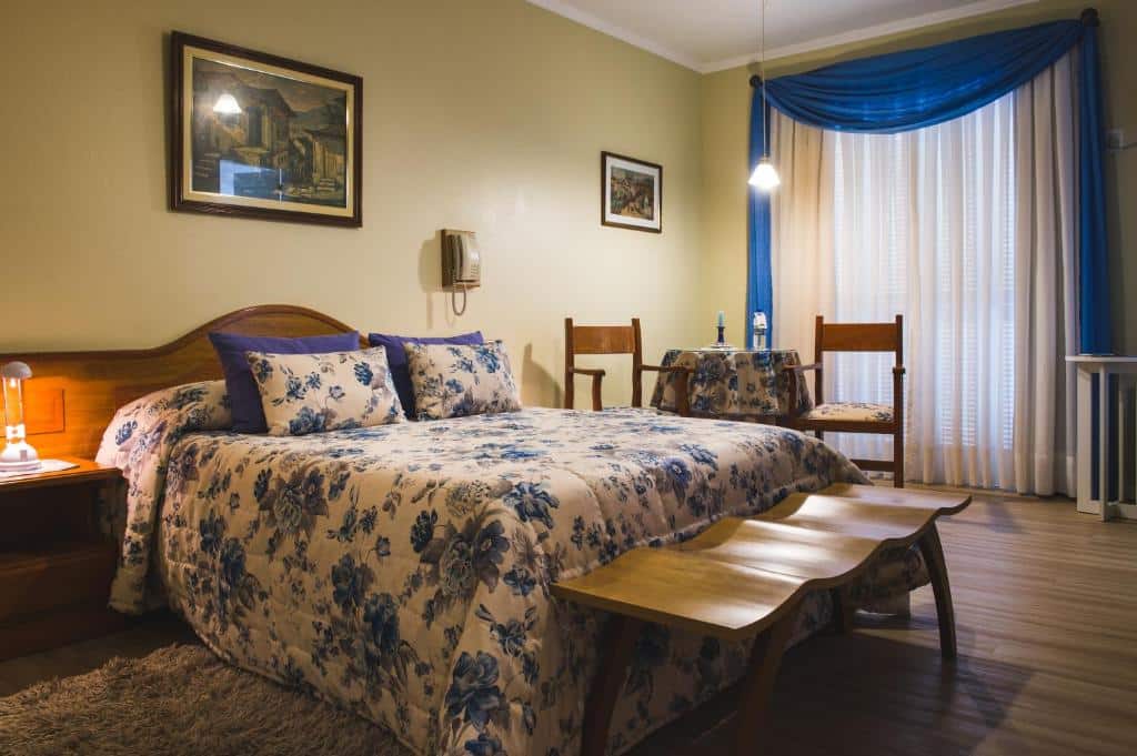 Uma cama bem arrumada na Pousada da Neve com roupa de cama floral azul em um quarto arrumado com móveis de madeira e cortinas azuis.