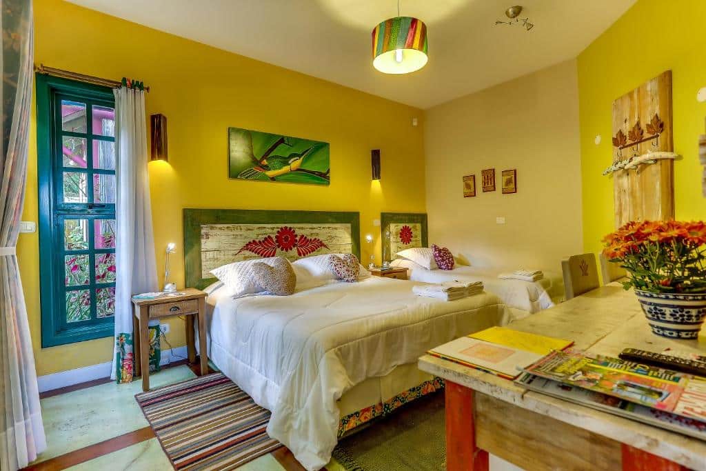 Um quarto aconchegante da Villa do Arquiteto com duas camas, paredes amarelas, decoração colorida, uma janela, mesas de cabeceira e uma cômoda com revistas.