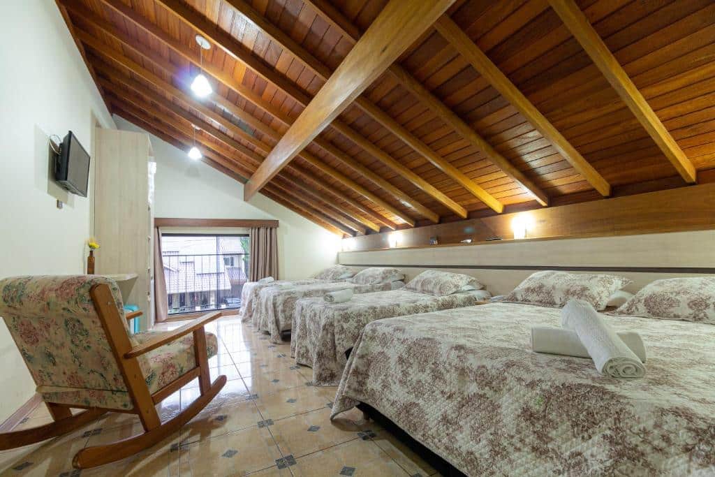 Quarto da Pousada Park Haus com quatro camas de casal, uma cadeira de balanço floral, TV na parede e teto inclinado de madeira.