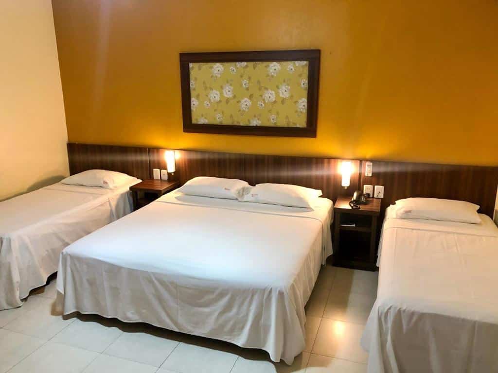 Quarto da Pousada Serrana com uma cama king-size e duas camas de solteiro, roupa de cama branca, mesas de cabeceira e uma parede amarela com obras de arte.