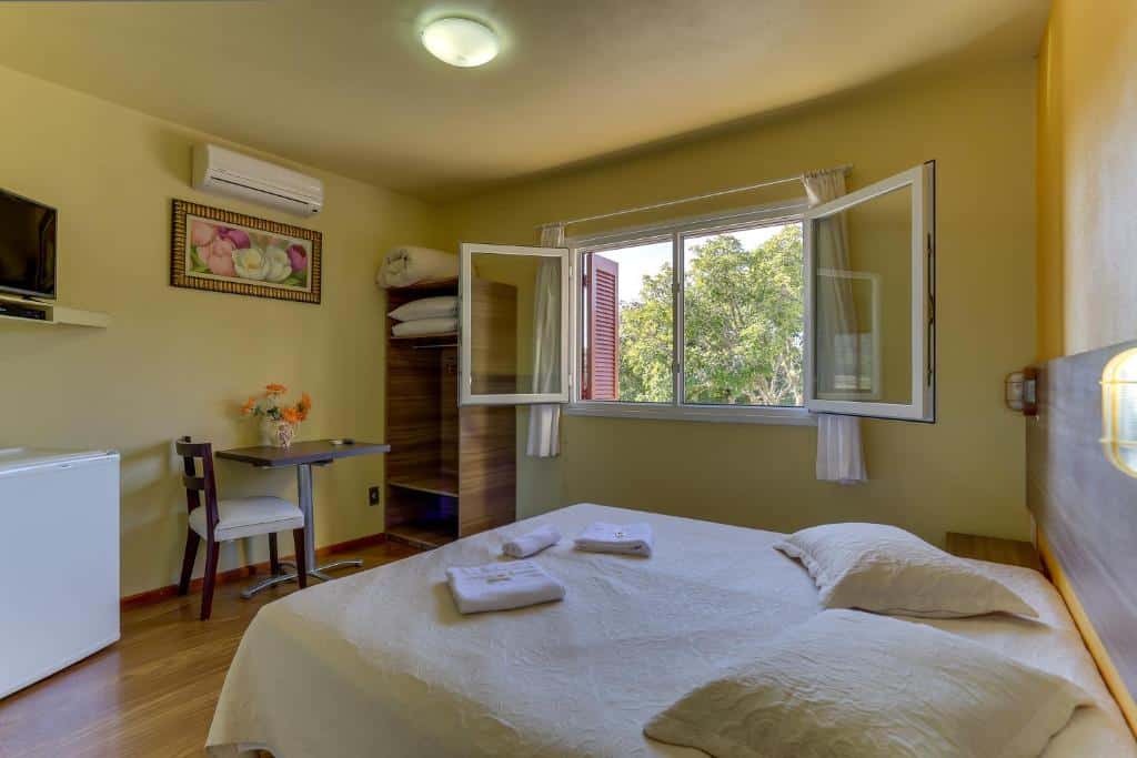 Uma cama bem arrumada em um quarto simples da Pousada Tio Mica com janela aberta, pequena escrivaninha, TV, frigobar e piso de madeira.